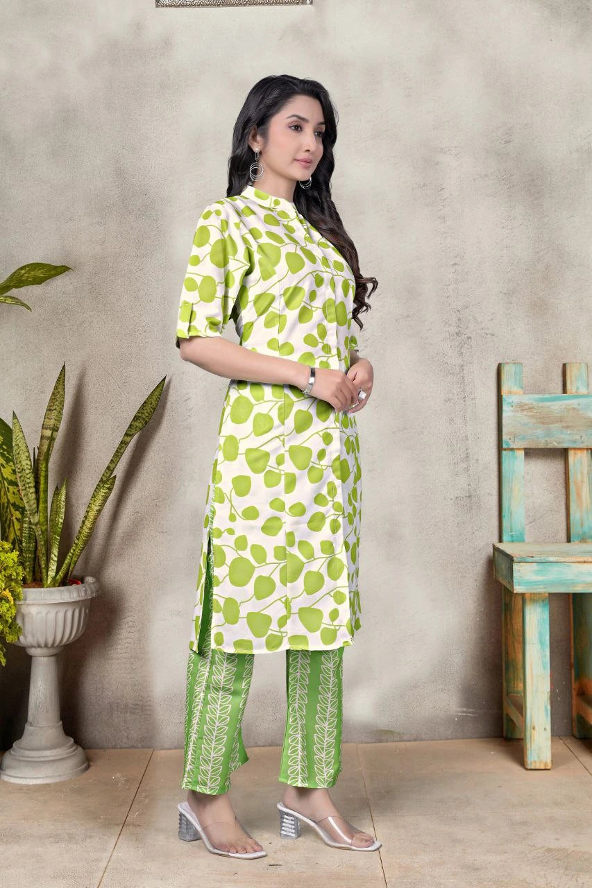 Woman wearing a green and white patterned outfit standing against a gray wall with a plant and wooden chair in the background.