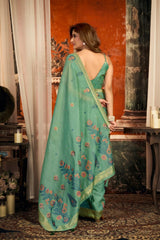 Woman in a green saree with floral patterns standing in a room with decorative elements.