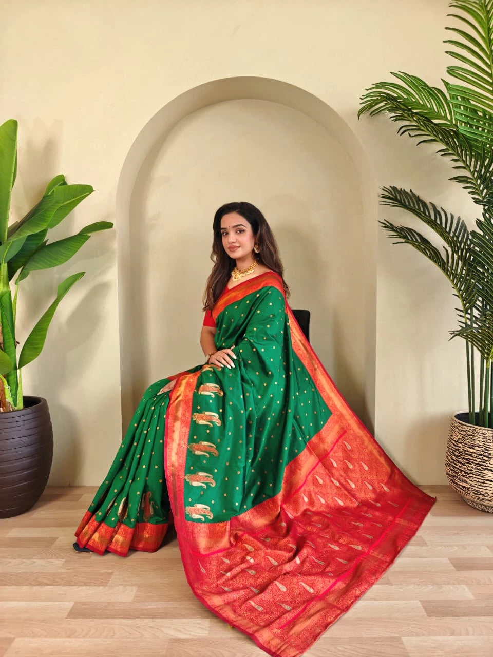 Woman in a green and red saree sitting indoors with plants around