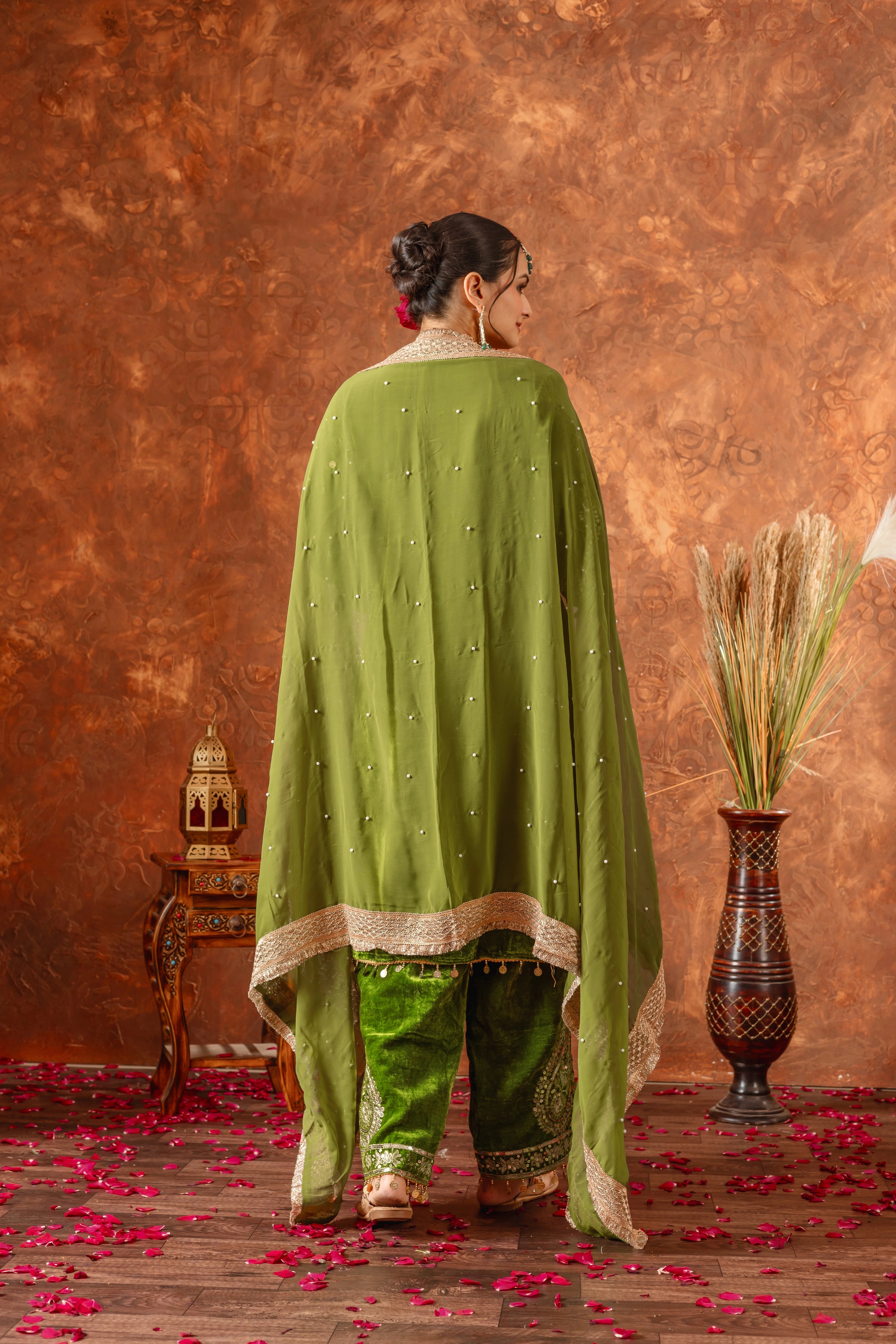 Woman in a green traditional outfit standing against a textured brown wall with decorative elements.