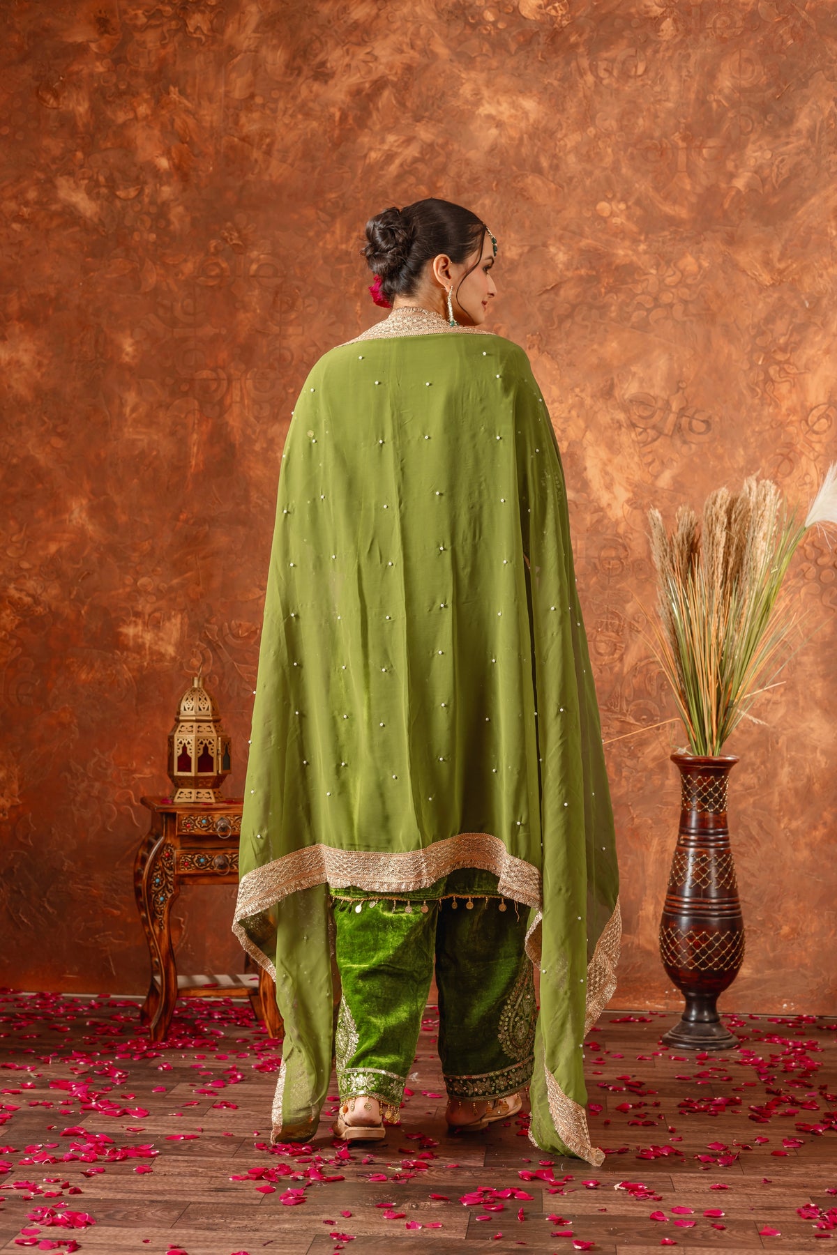 Woman in a green traditional outfit standing against a textured brown wall with decorative elements.