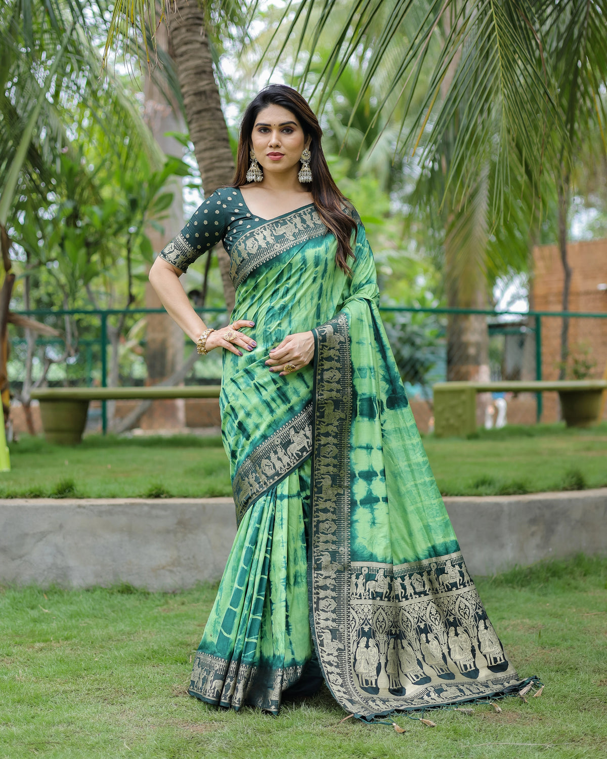Woman in a green saree with black patterns standing outdoors with trees in the background