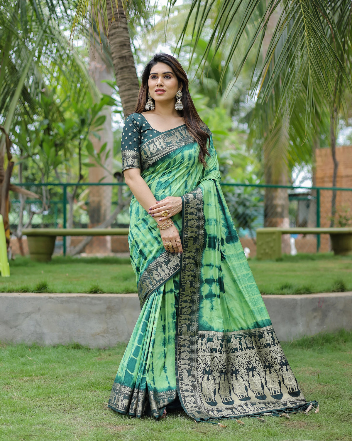 Woman in a green saree with black patterns standing outdoors with trees in the background