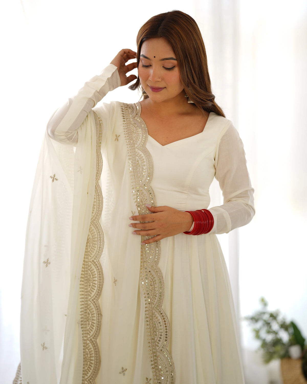 Woman wearing a white traditional outfit with a sheer dupatta, posing indoors.