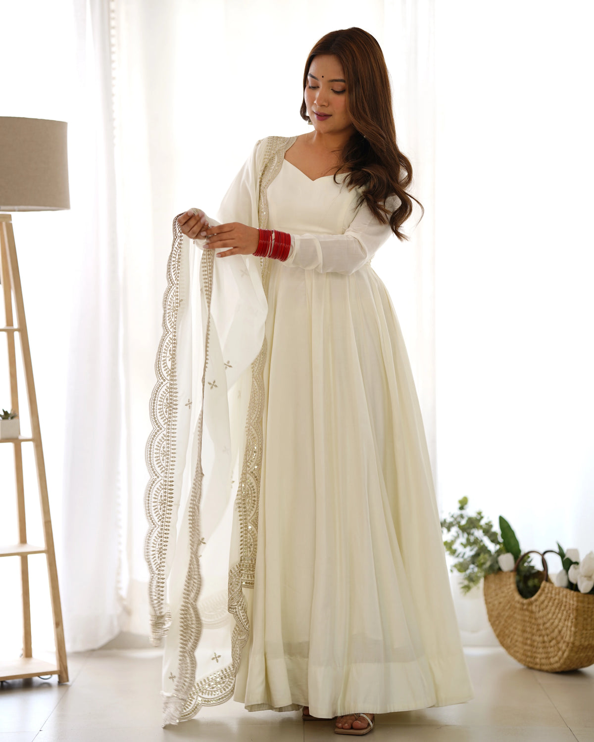 Woman in a long white dress with a sheer dupatta in a bright room.
