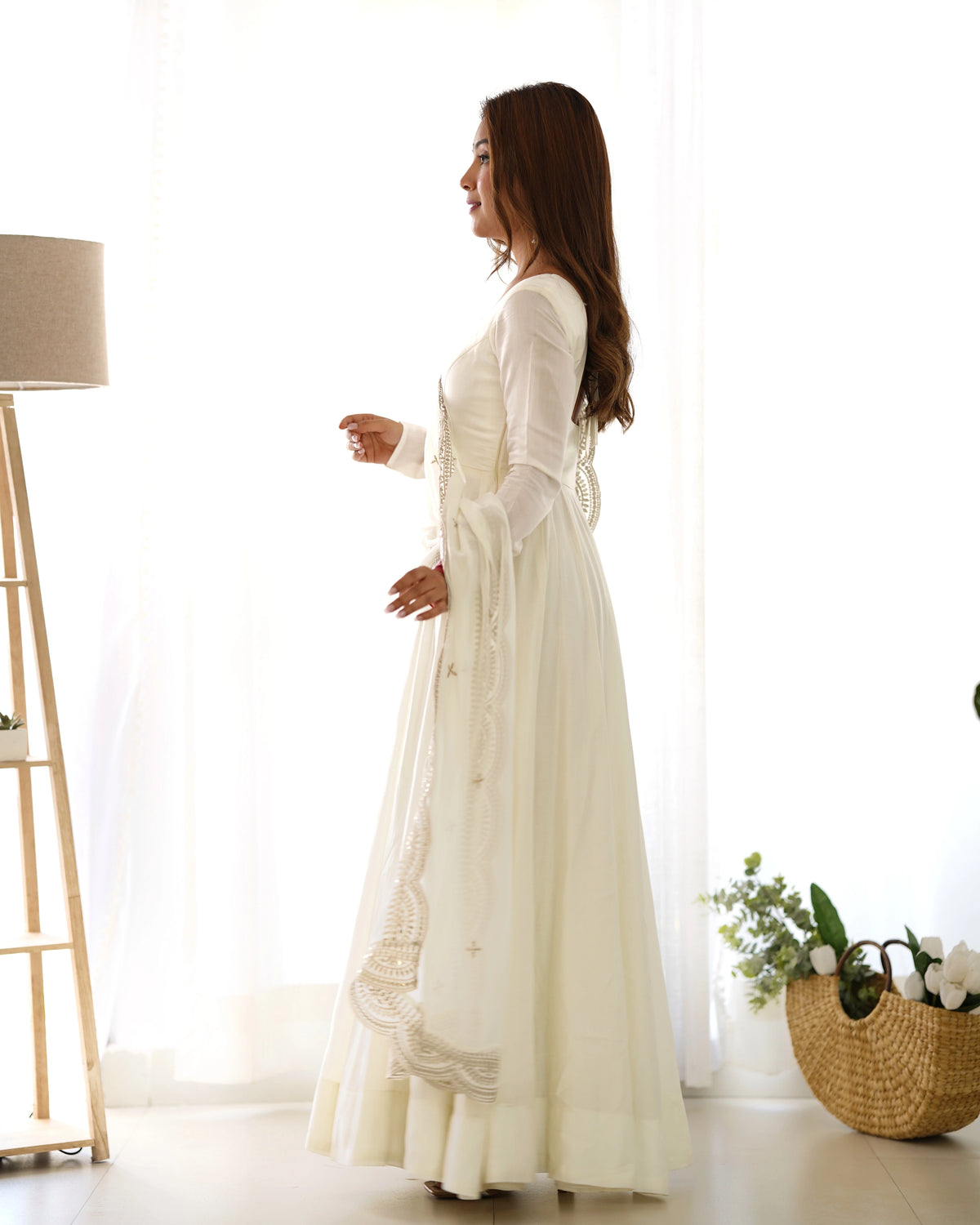 Woman in a long white dress standing in a bright room with a lamp and basket in the background.