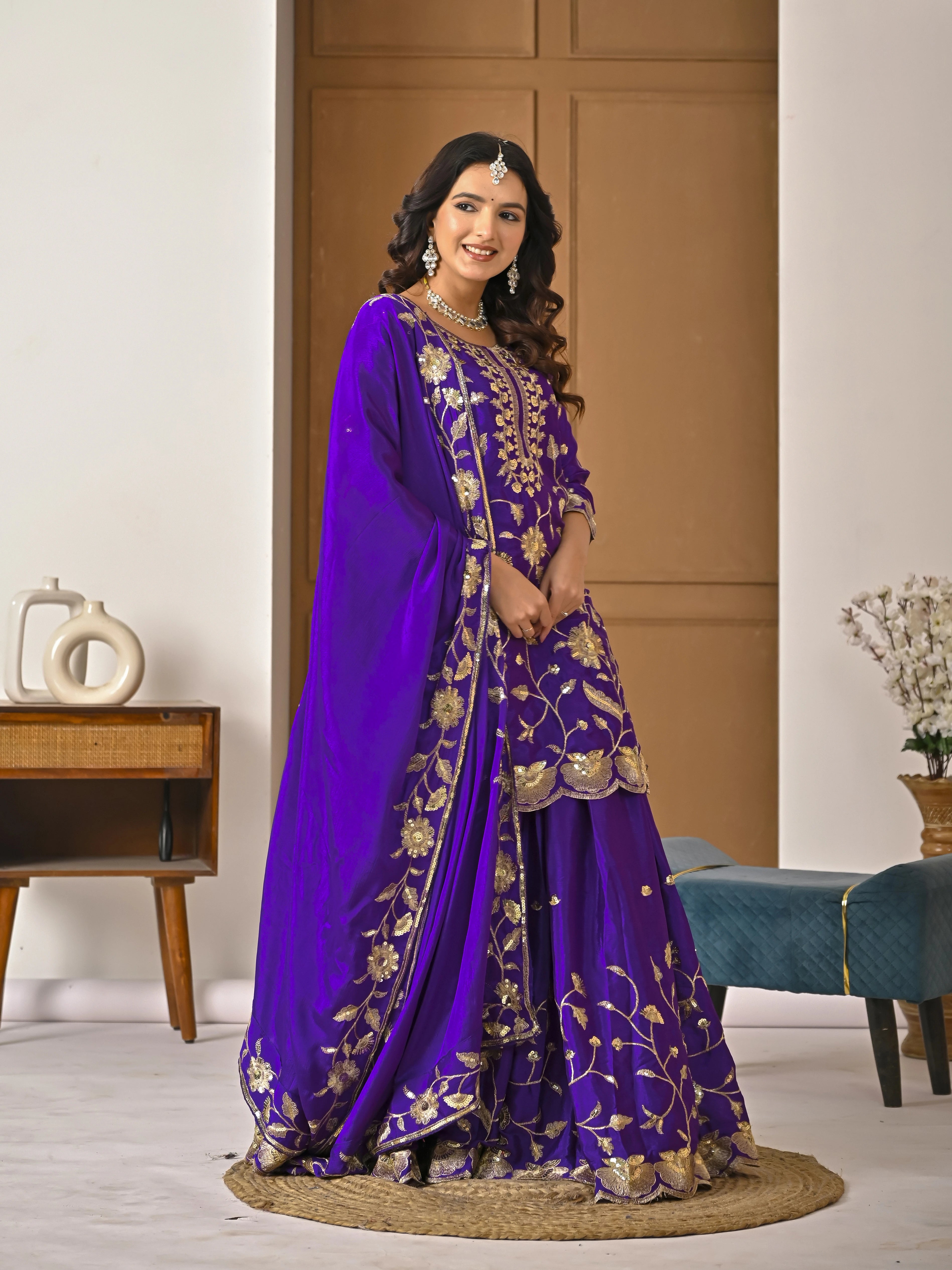 Woman wearing a purple embroidered traditional outfit in a room with a wooden cabinet and decorative items.