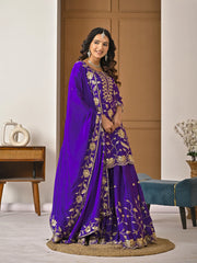 Woman wearing a purple embroidered traditional outfit in a room with a wooden cabinet and decorative items.