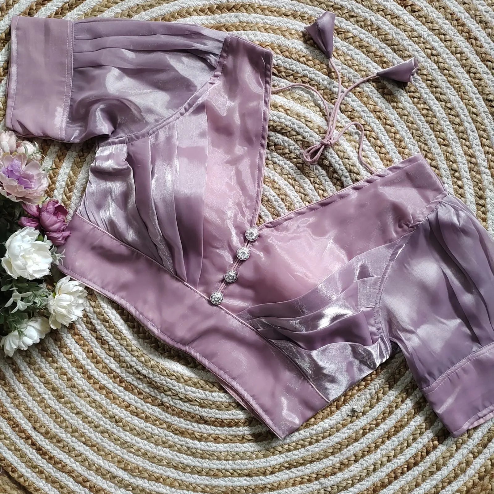 Lavender satin blouse with pleated detailing and four decorative silver buttons on the front, featuring elbow-length sleeves and tie-up tassels, displayed on a circular woven mat with white and purple flowers on the side