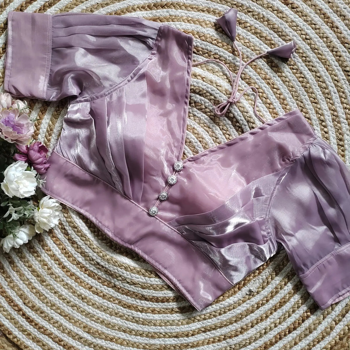 Lavender satin blouse with pleated detailing and four decorative silver buttons on the front, featuring elbow-length sleeves and tie-up tassels, displayed on a circular woven mat with white and purple flowers on the side