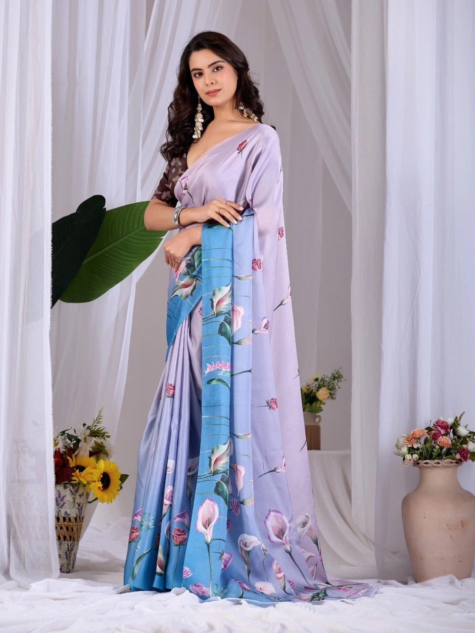 Woman wearing a light purple saree with floral patterns, standing against a white curtain background.