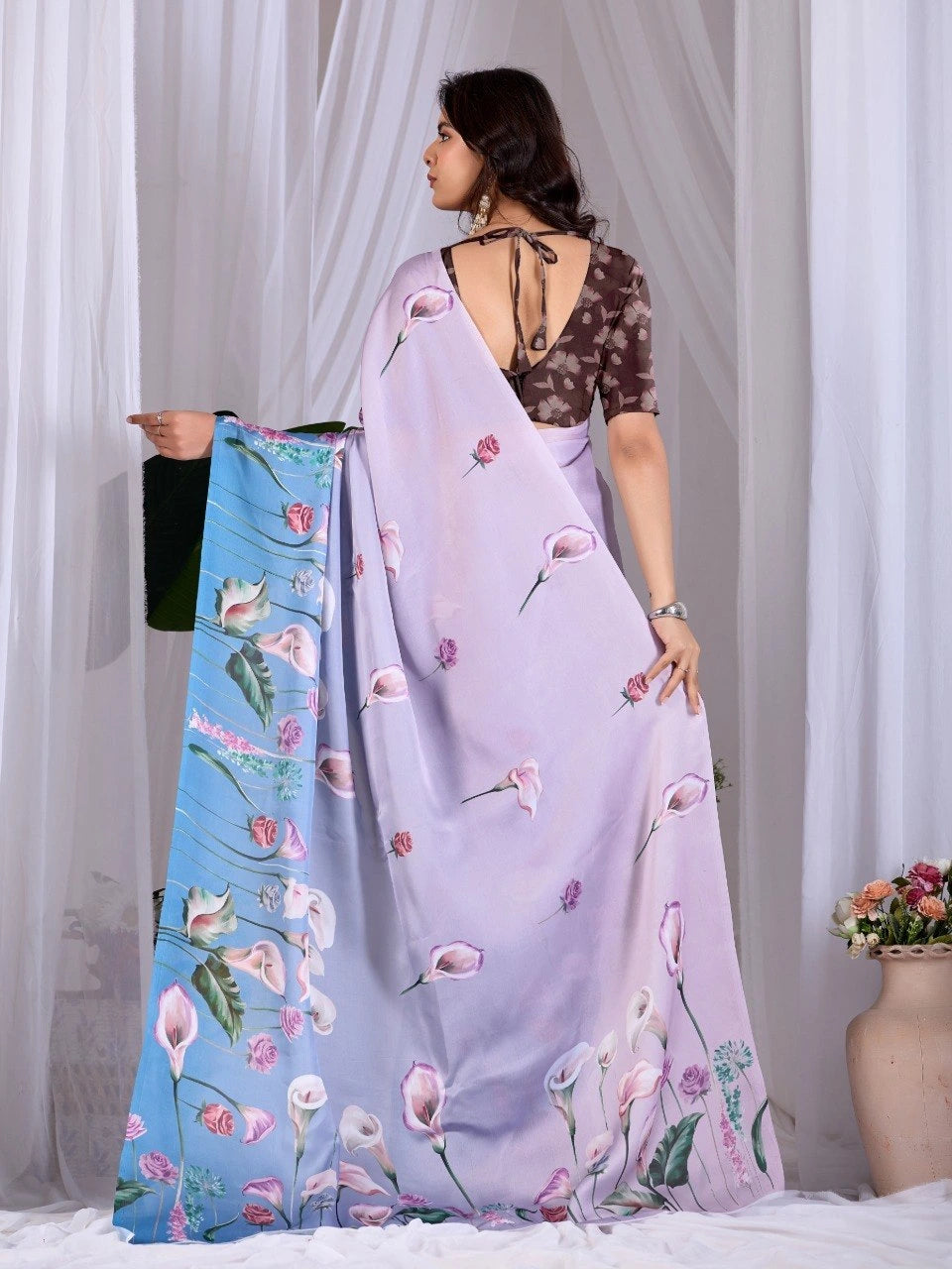 Woman holding a floral saree with a plain blue saree, against a white curtain background.