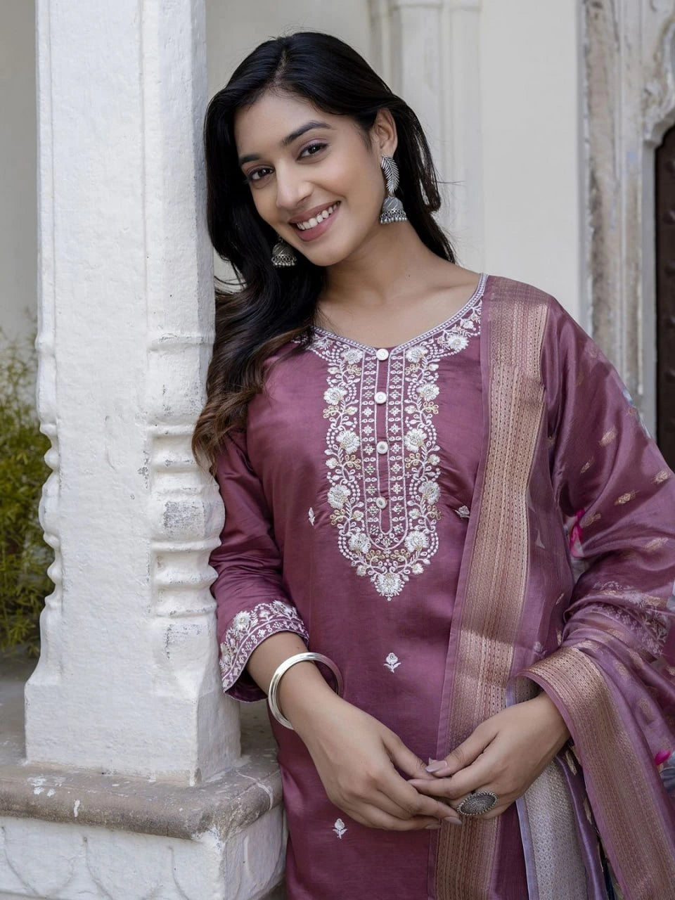 Woman wearing a traditional embroidered purple outfit standing against a white architectural background.