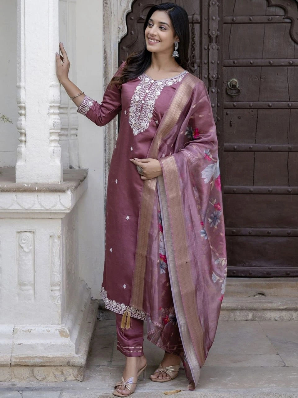 Woman in a traditional maroon outfit with floral patterns standing against a stone wall.
