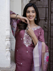 Woman in a maroon traditional outfit with white embroidery standing against a textured wall.