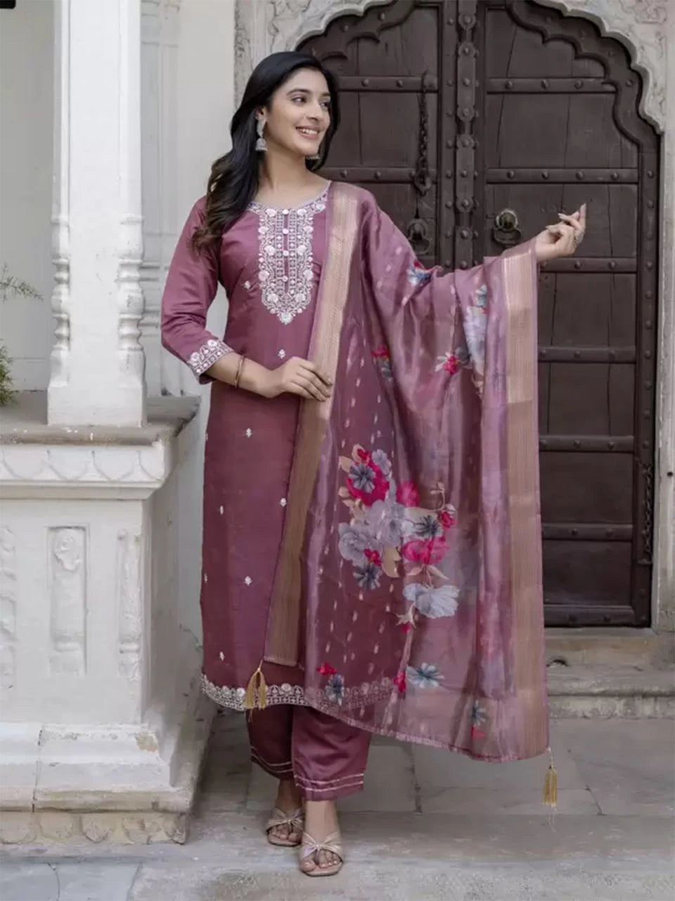 Woman in traditional maroon outfit with floral dupatta standing in front of a decorative door.