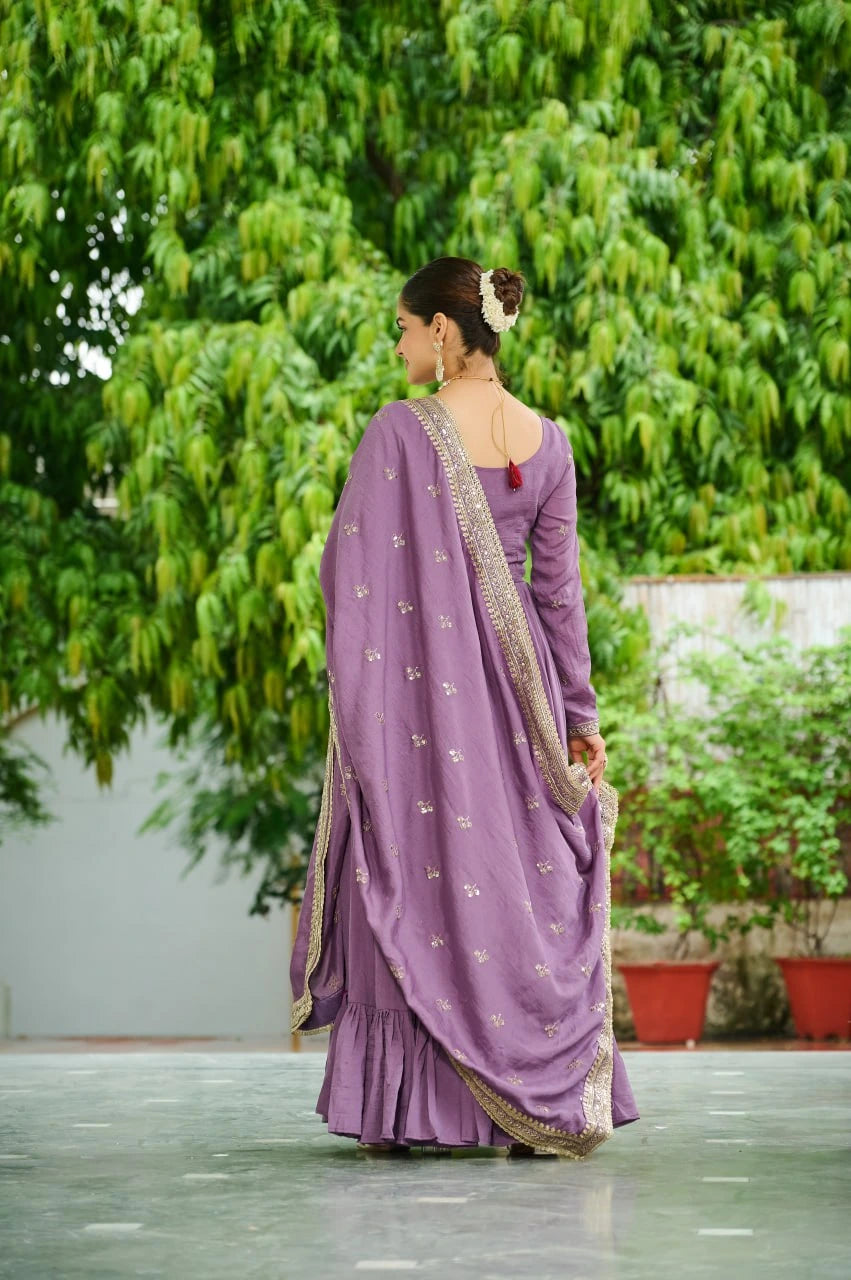 Woman in a purple saree with a gold border standing in front of green foliage.