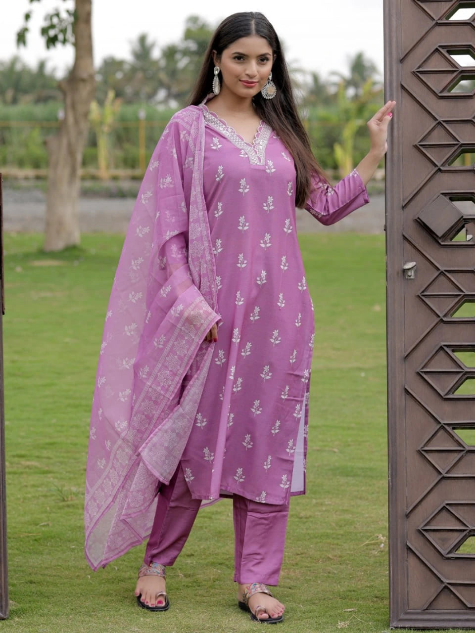 Woman in a purple traditional outfit standing outdoors near a decorative gate.