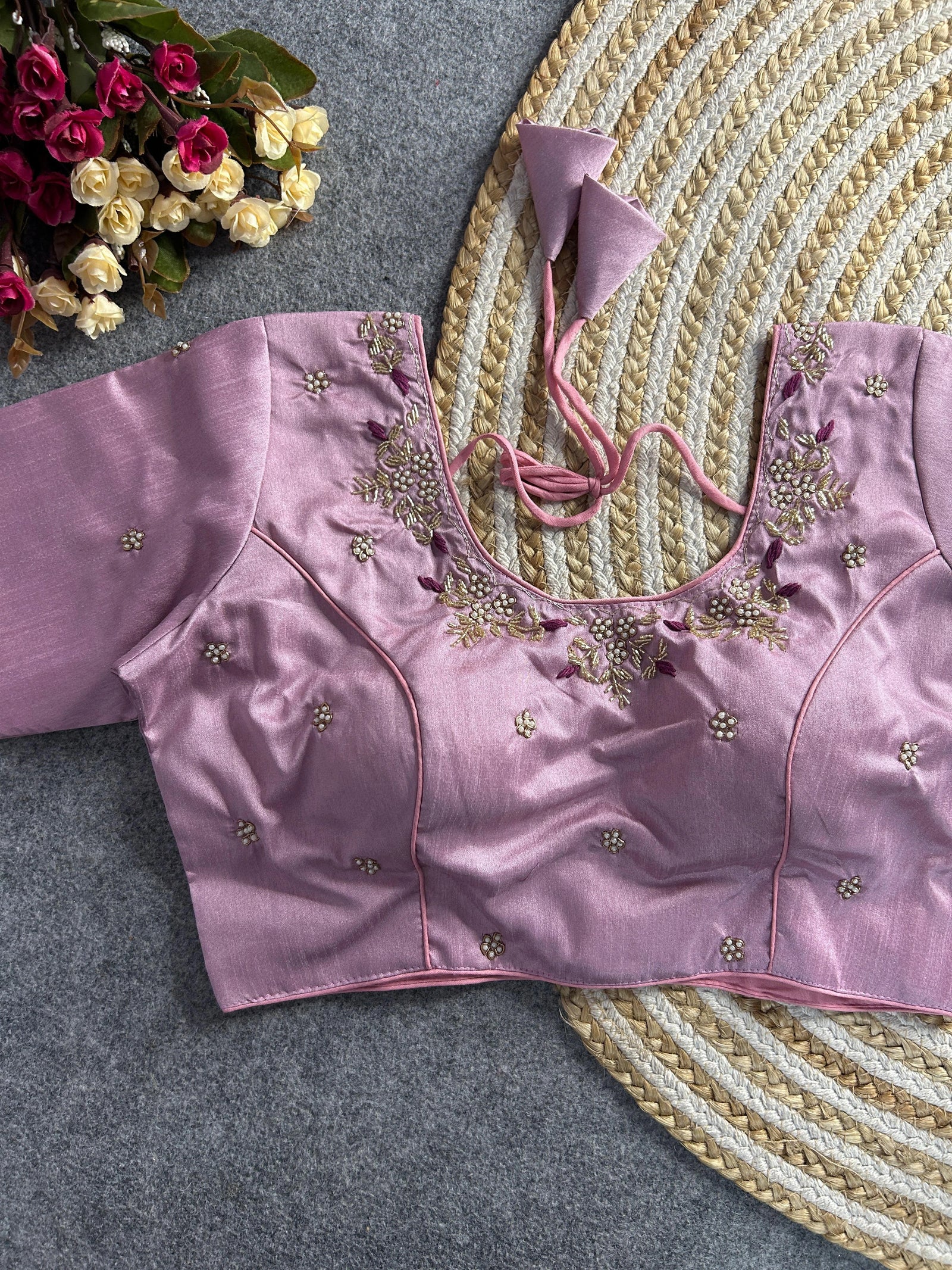 The front of the lavender silk blouse shows a curved V-neckline with matching pearl and gold floral embroidery. The bodice is structured with princess-cut seams and adorned with scattered pearl floral motifs. The blouse is laid flat on a grey textured surface next to a decorative jute mat and a bouquet of roses.