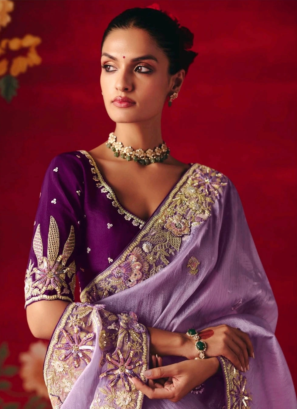 Woman wearing a traditional purple saree with gold embroidery against a red background