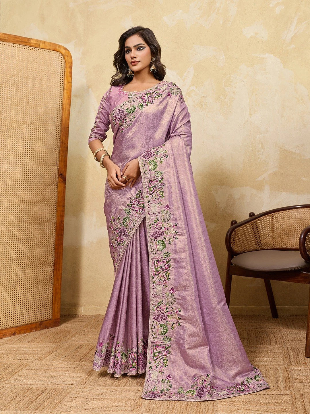 Woman wearing a lavender saree with floral patterns in a room with a textured wall and wooden chair.