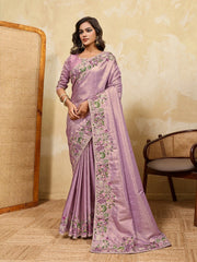 Woman wearing a lavender saree with floral patterns in a room with a textured wall and wooden chair.