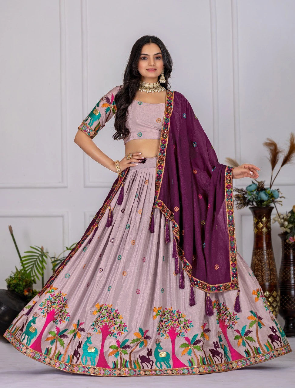 Woman wearing a traditional outfit with a pink lehenga and purple dupatta against a white wall.