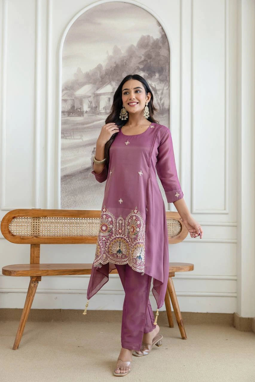Woman in a purple traditional outfit standing in a room with a wooden bench and decorative wall.