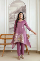 Woman wearing a purple embroidered outfit standing in a room with a wooden bench and decorative wall.