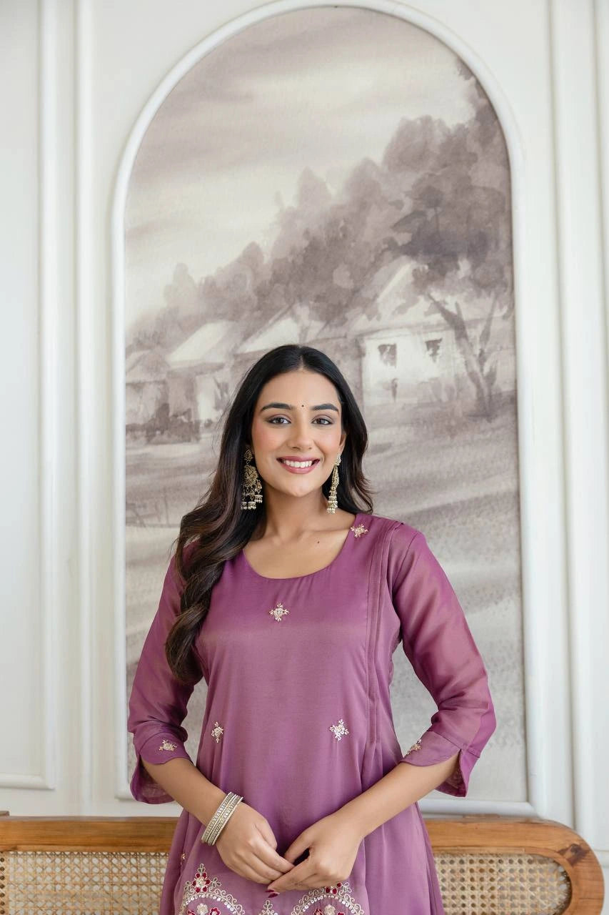 Woman in a purple outfit standing in front of a decorative wall with a landscape painting.