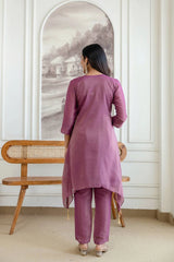 Woman in a purple outfit standing in a room with a wooden chair and framed picture on the wall.