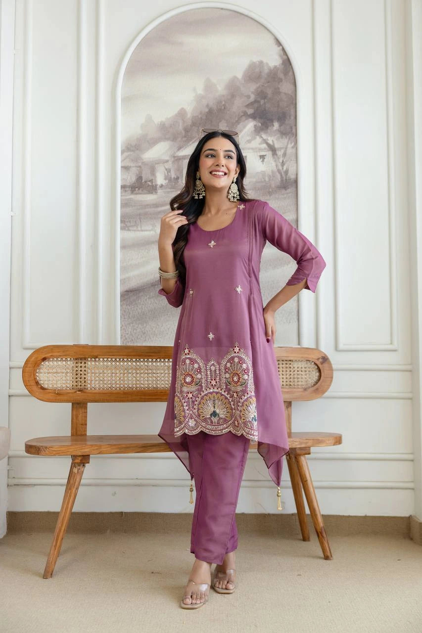 Woman in a purple outfit with intricate patterns standing next to a wooden bench in a room with white walls and a decorative mirror.