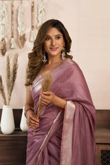 Woman in a pink saree holding a plant, standing against a decorative wall.