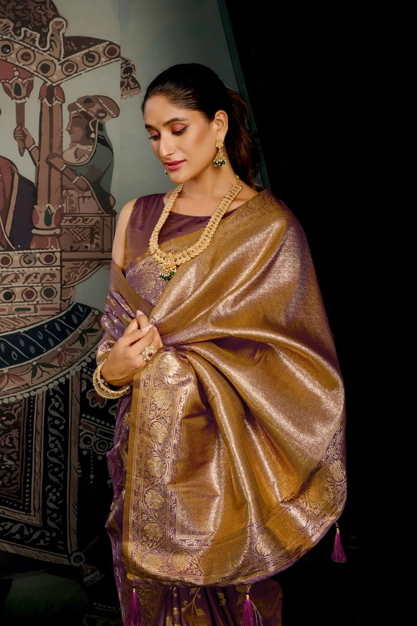 Woman wearing a traditional saree with a gold and purple color scheme, standing against a dark background.