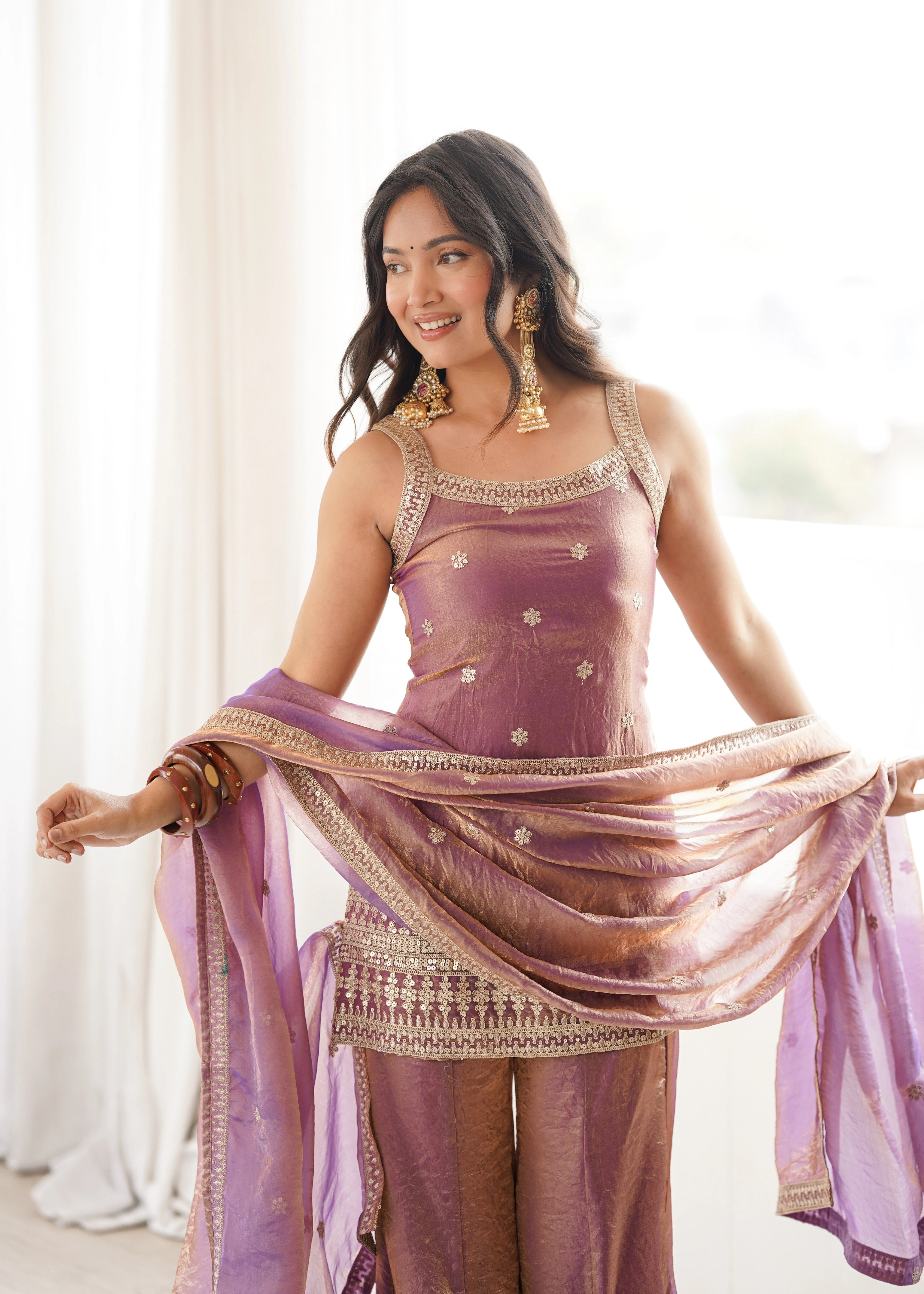 Woman in a traditional outfit with a purple dupatta, standing indoors.