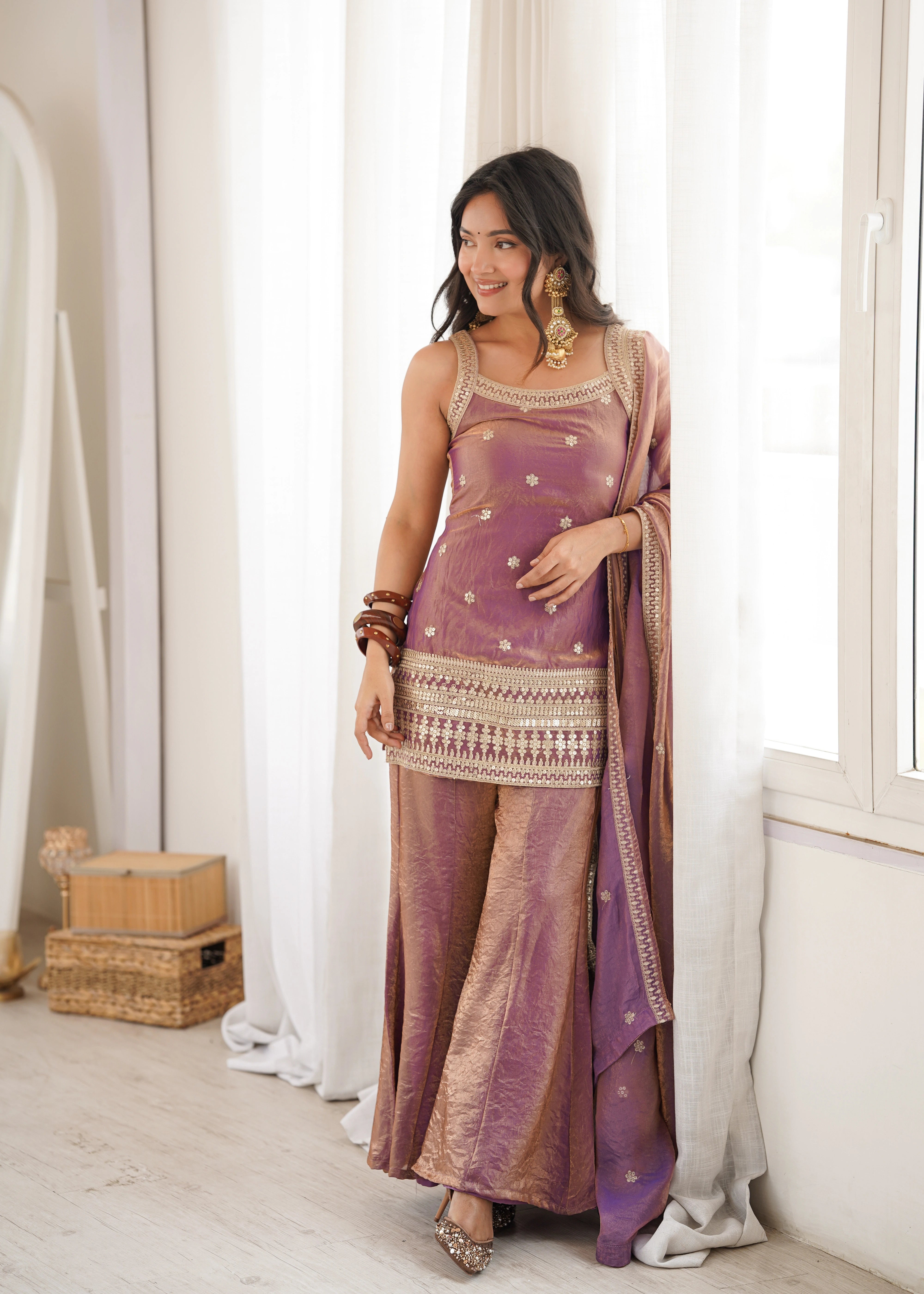 Woman in a traditional outfit standing in a room with white curtains and wooden boxes.