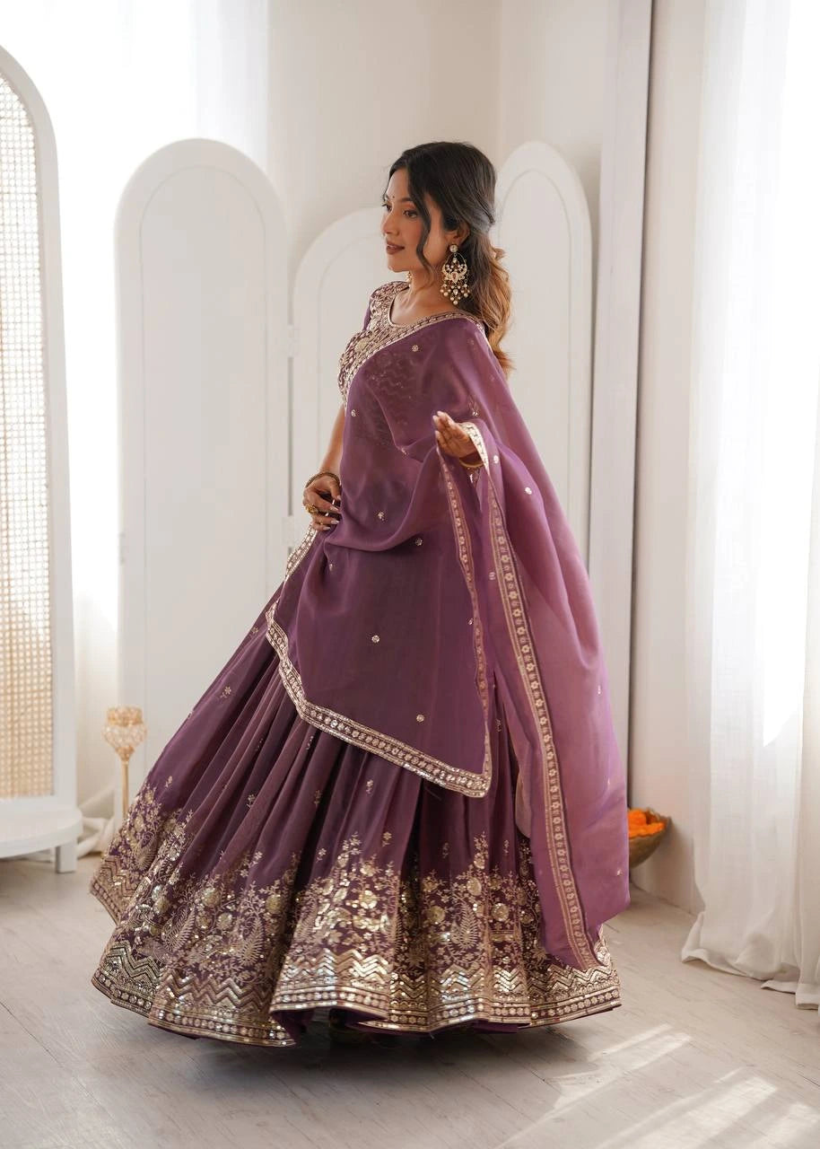 Woman in a purple traditional outfit with gold embroidery standing in a bright room.