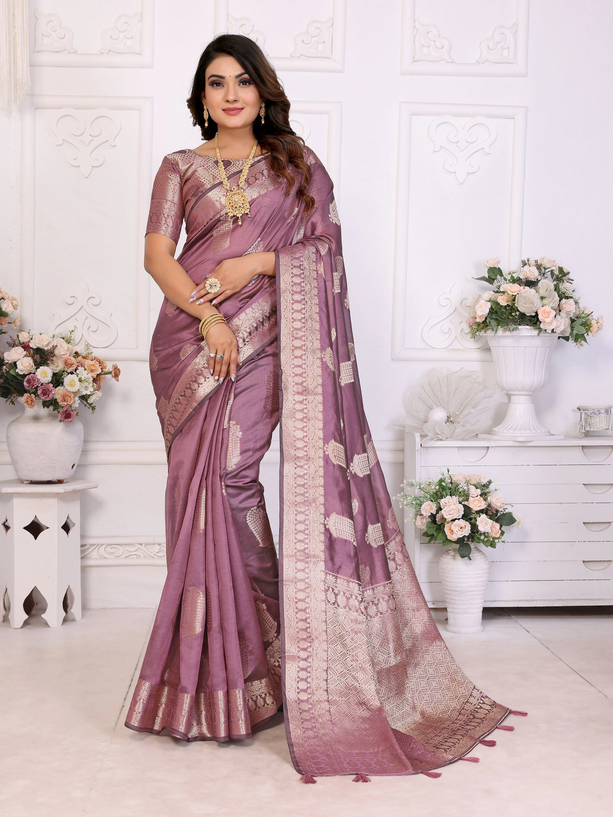 Woman wearing a purple saree with gold patterns in a decorated room.