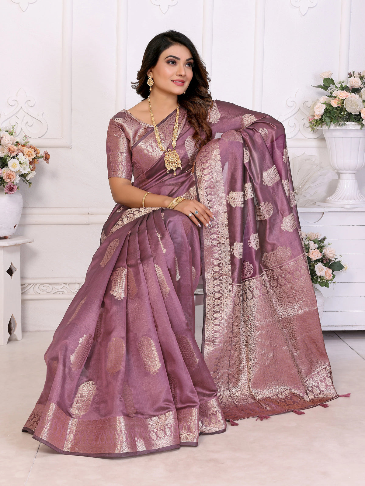 Woman wearing a purple saree with gold patterns in a decorated room.