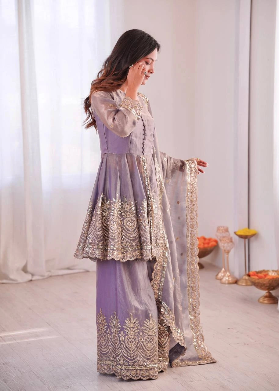 Woman in a traditional outfit with intricate designs, standing in a room with white curtains and decorative items.