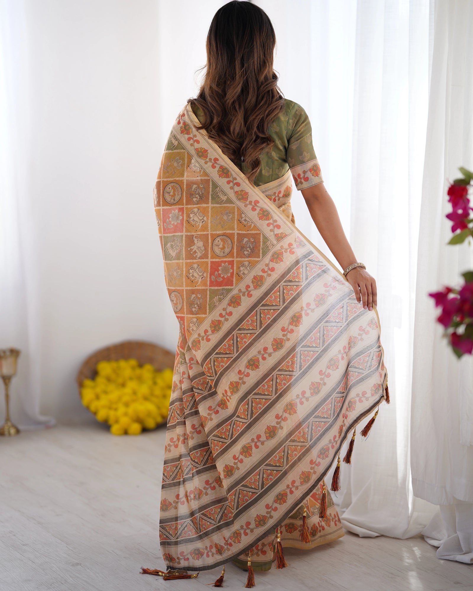 Woman wearing a patterned saree in a bright room with white curtains and flowers.