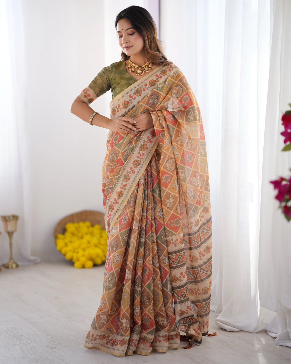 Woman wearing a patterned saree in a bright room with white curtains and flowers.