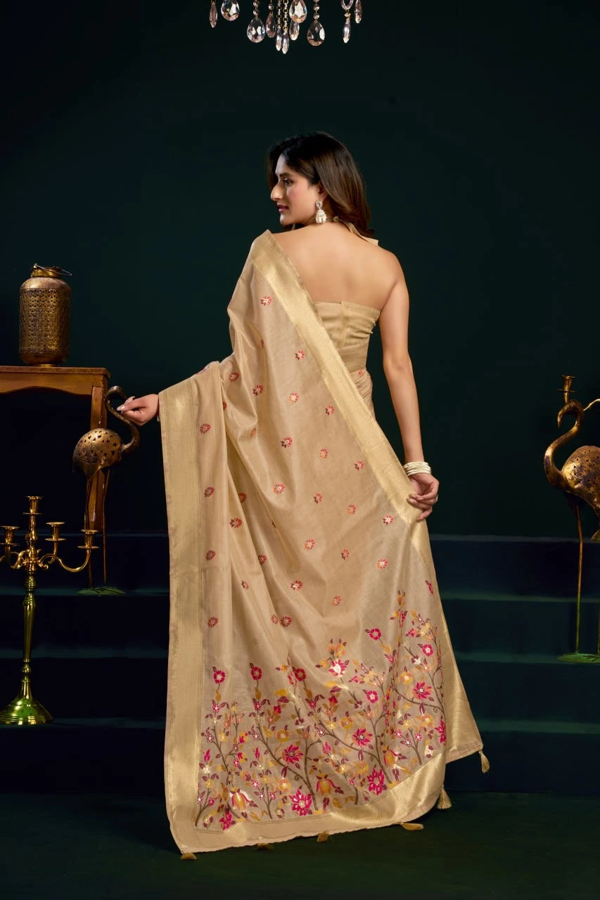 Woman in a beige saree with floral patterns standing in a dark room.