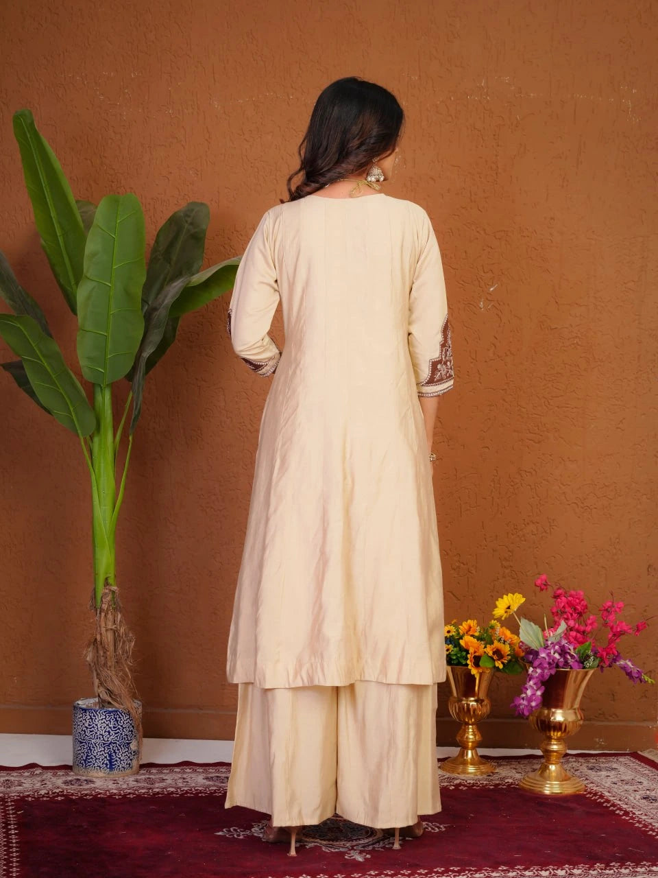 Woman in a beige traditional outfit standing in front of a brown wall with decorative elements.