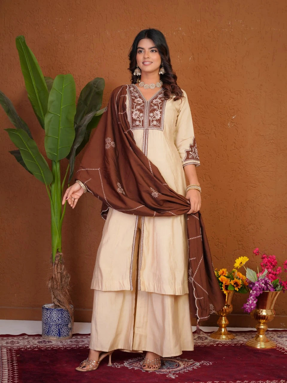 Woman in a traditional outfit with a brown dupatta standing against a brown wall with decorative elements.