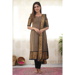 Woman wearing a brown and black traditional outfit with intricate designs, standing against a white wall.