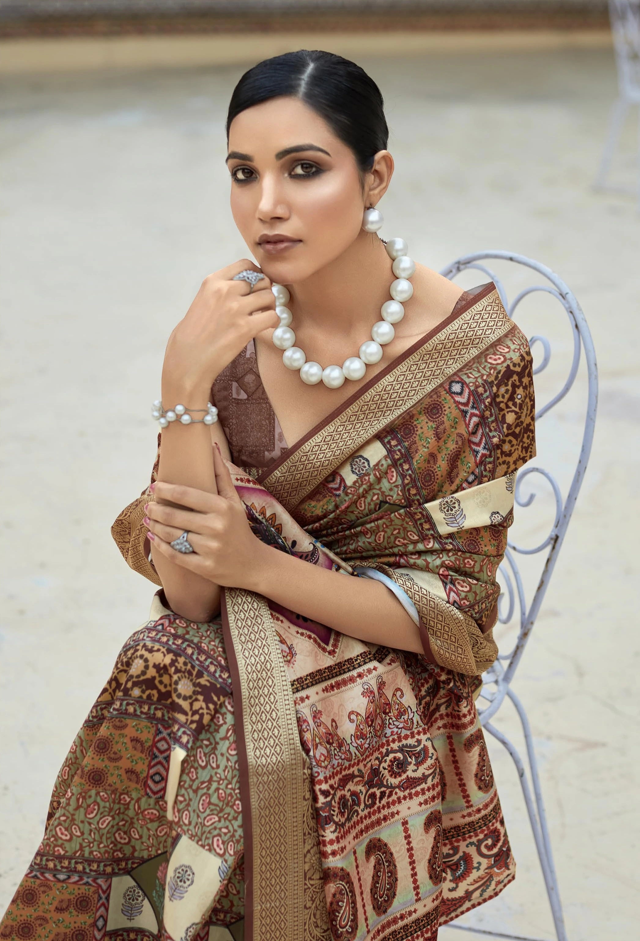 Woman wearing a patterned saree with jewelry, sitting on a chair.