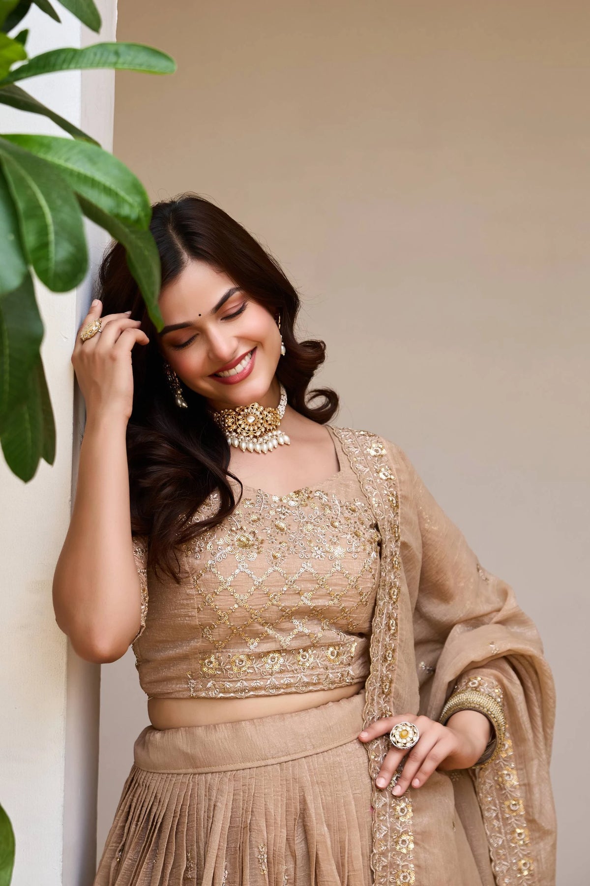 Woman in traditional beige outfit with jewelry, standing next to a plant.