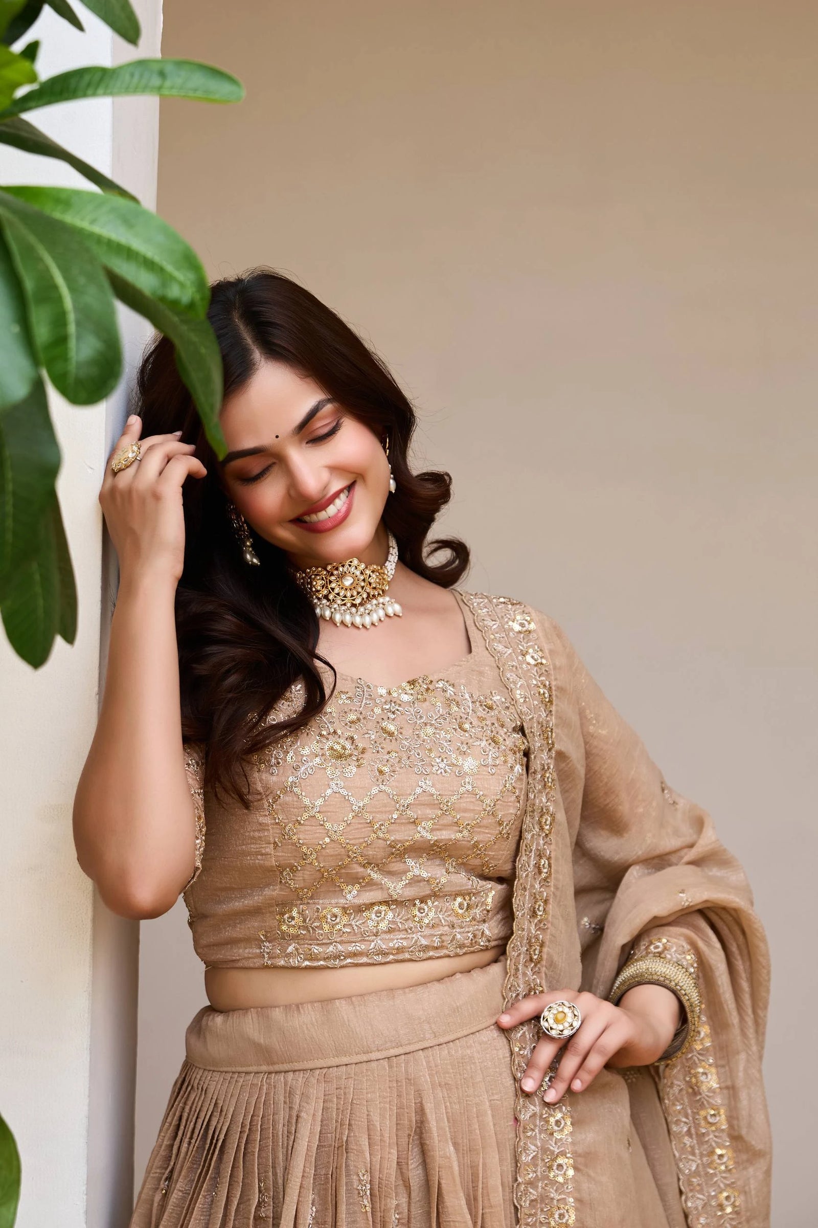 Woman in traditional beige outfit with jewelry, standing next to a plant.