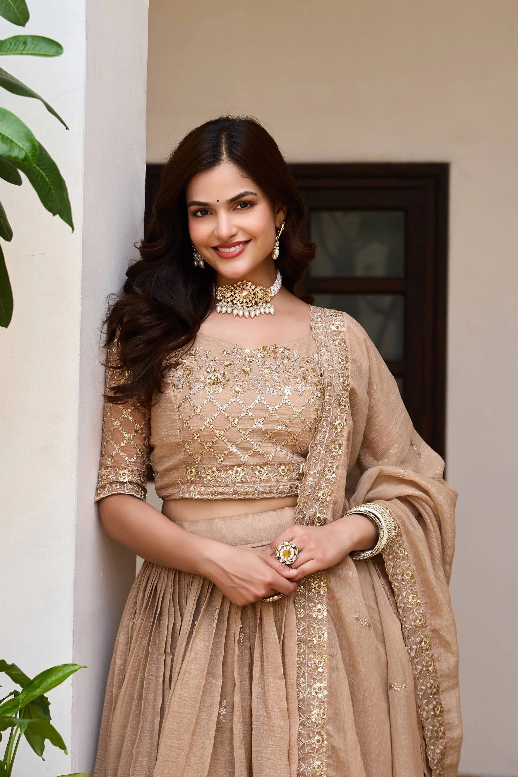Woman in a beige embroidered outfit standing indoors with plants in the background